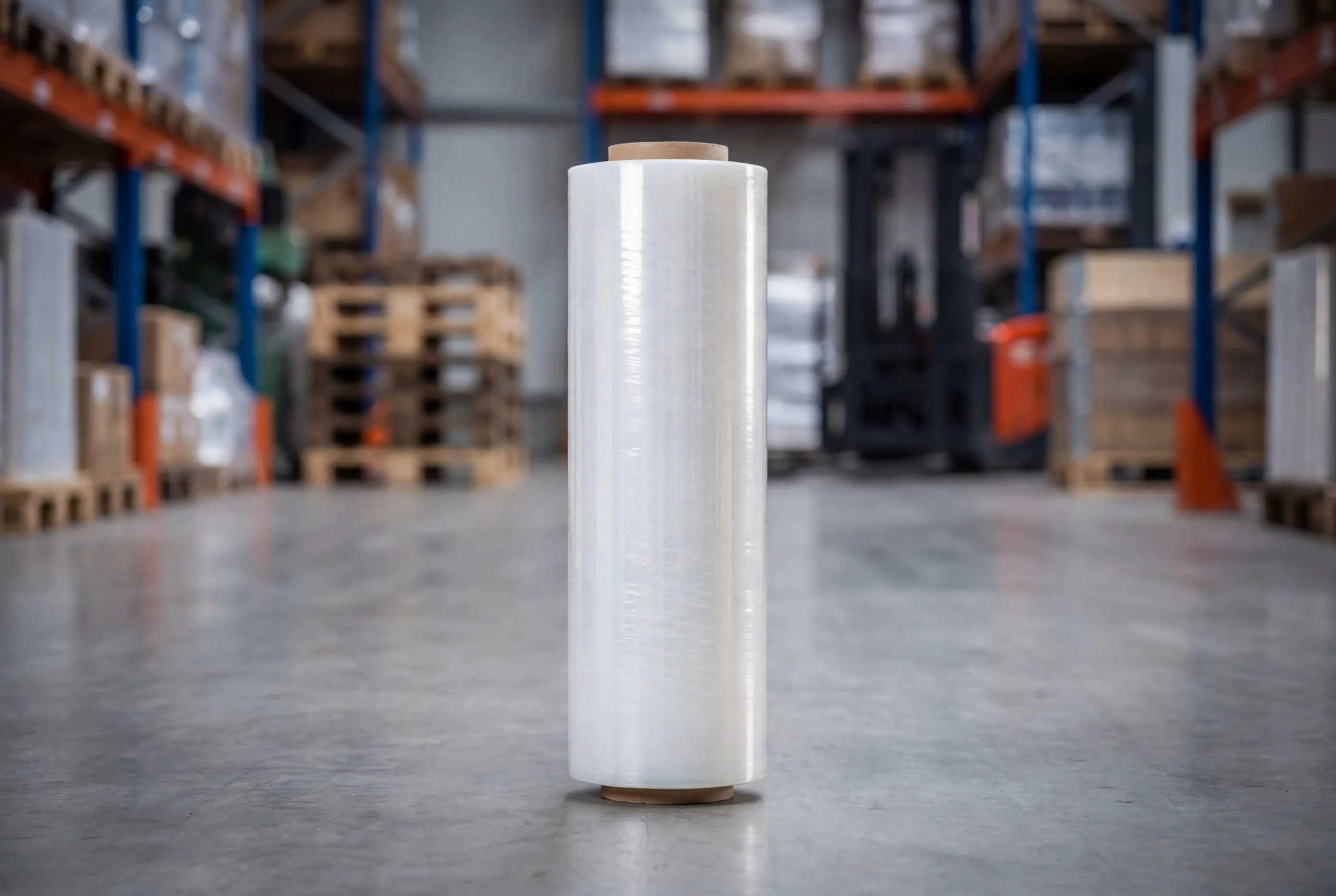 Un gran rollo de film estirable de plástico transparente está en posición vertical en el suelo de un almacén, con estanterías, palés y cajas visibles en el fondo borroso.