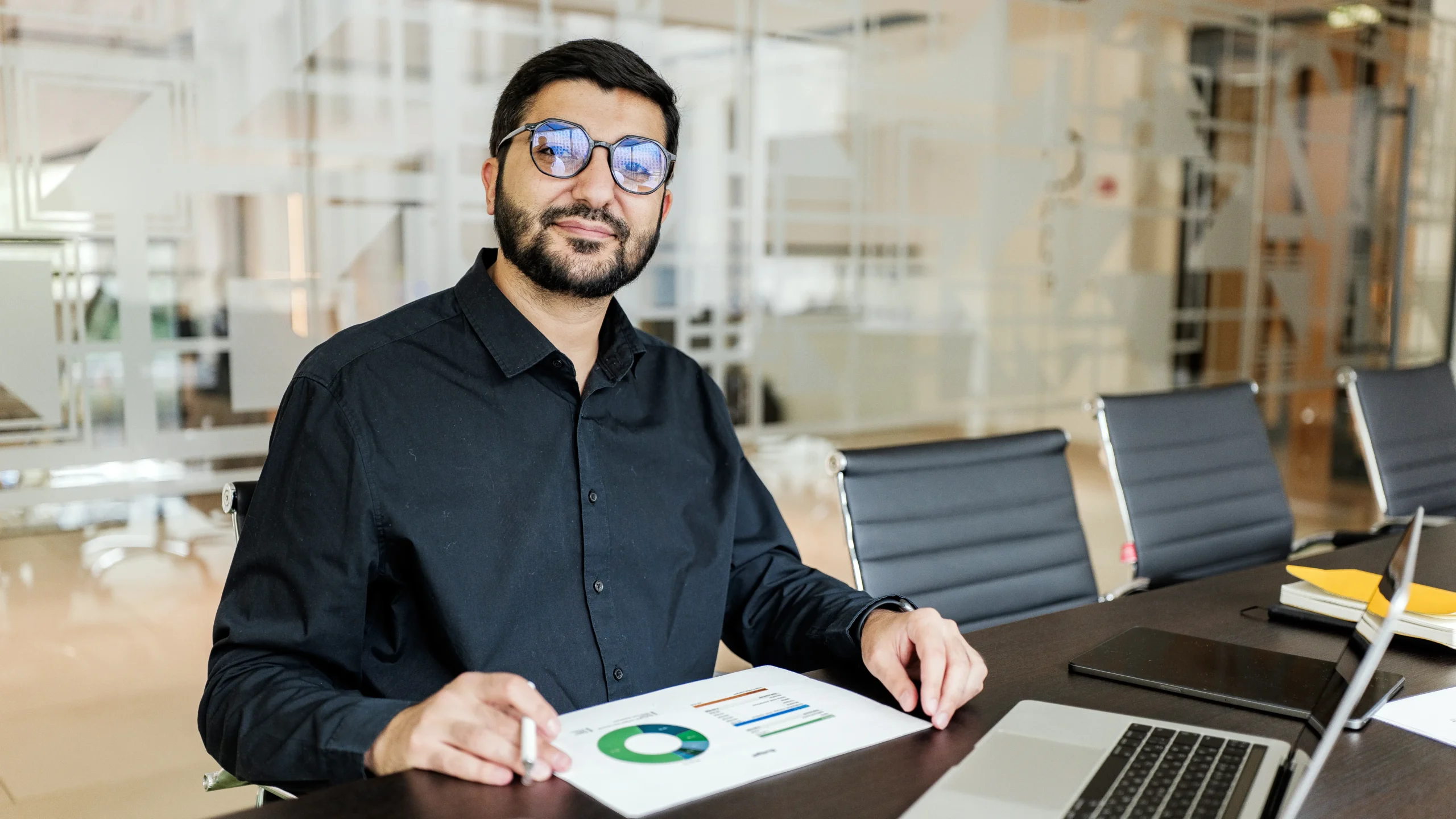 Hombre con lentes sentado en mesa de sala de juntas, señalando un documento con gráficas de resultados junto a una laptop