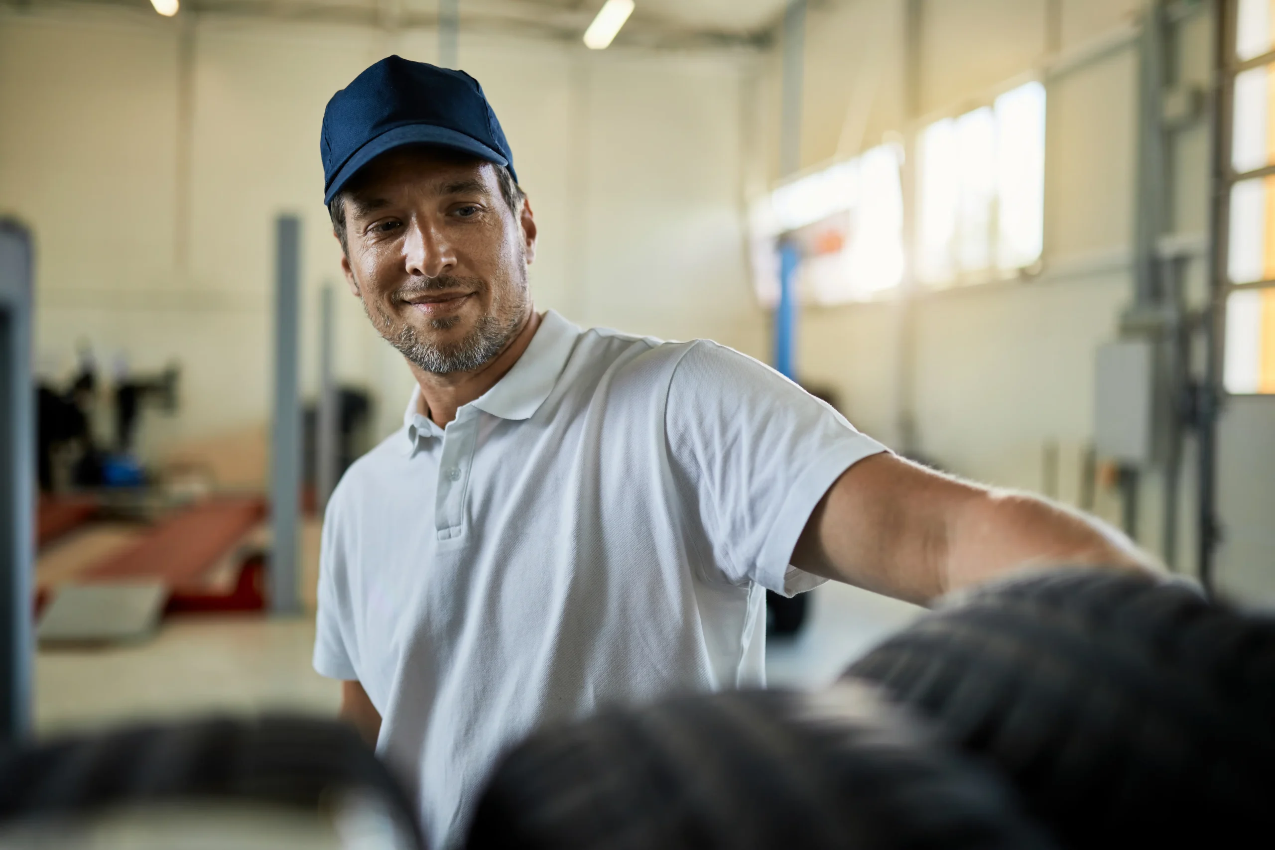 Hombre con gorra y playera polo blanca de pie en taller automotriz, con llantas apiladas en primer plano