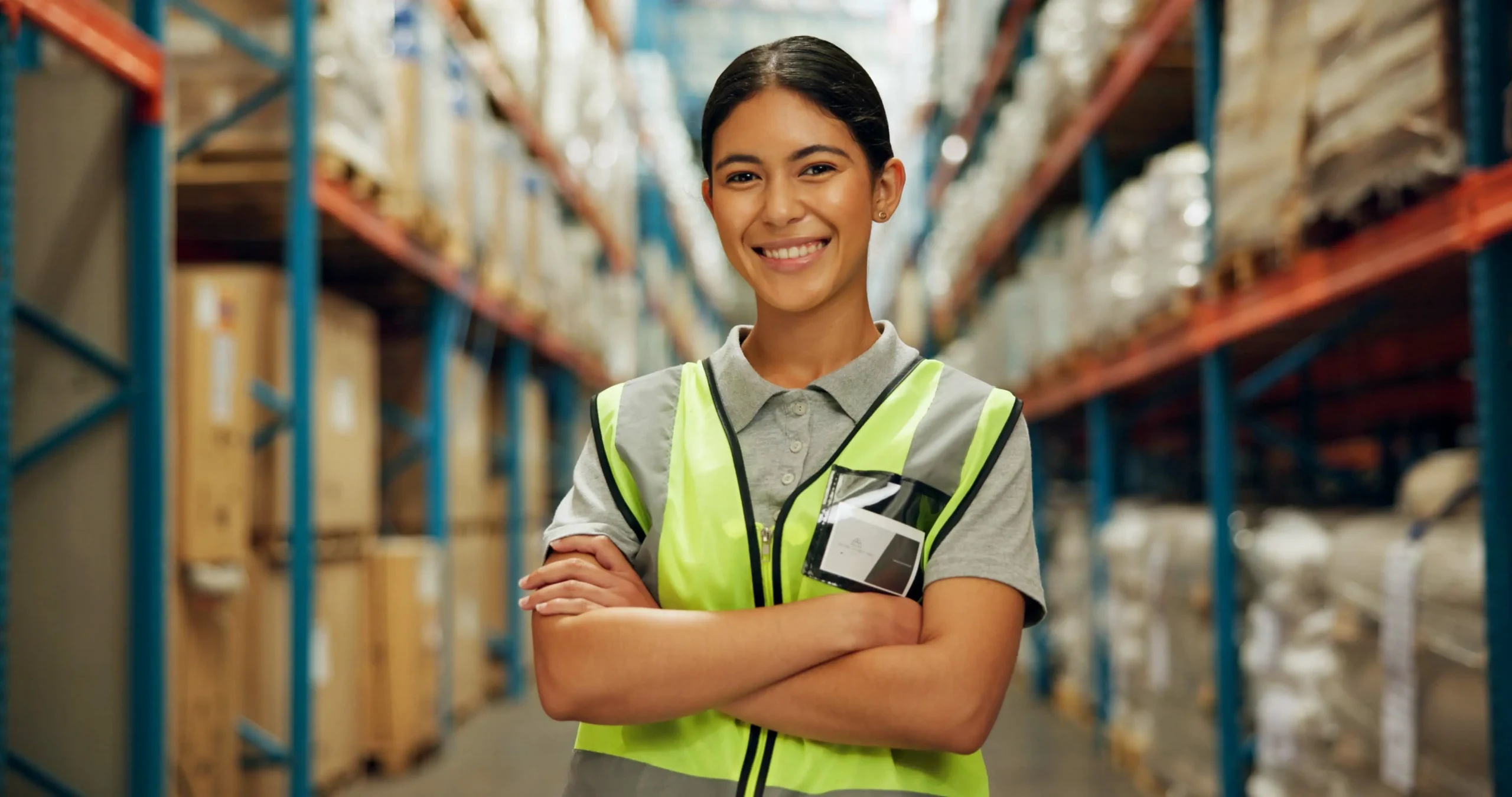 Trabajadora de almacén con chaleco de seguridad reflectante, de pie entre pasillos de racks con tarimas y mercancía embalada