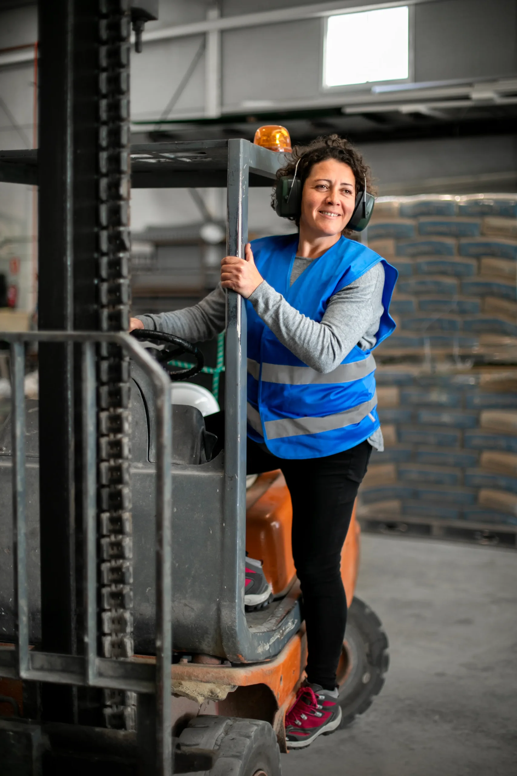 Mujer con chaleco azul y orejeras de protección subiendo a un montacargas, con sacos apilados de fondo en bodega industrial