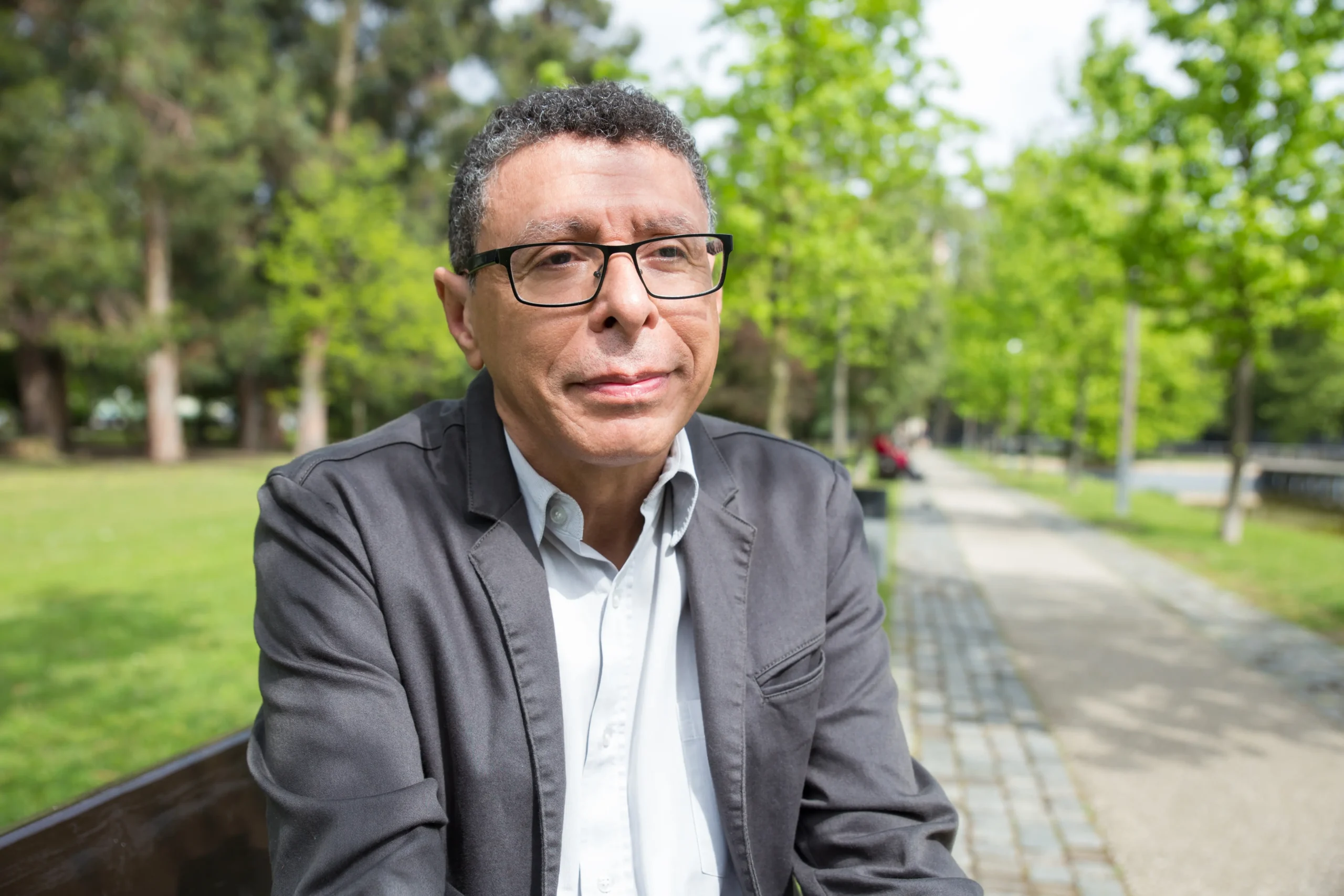 Hombre de mediana edad con lentes y saco gris, sentado en una banca de parque con vegetación de fondo
