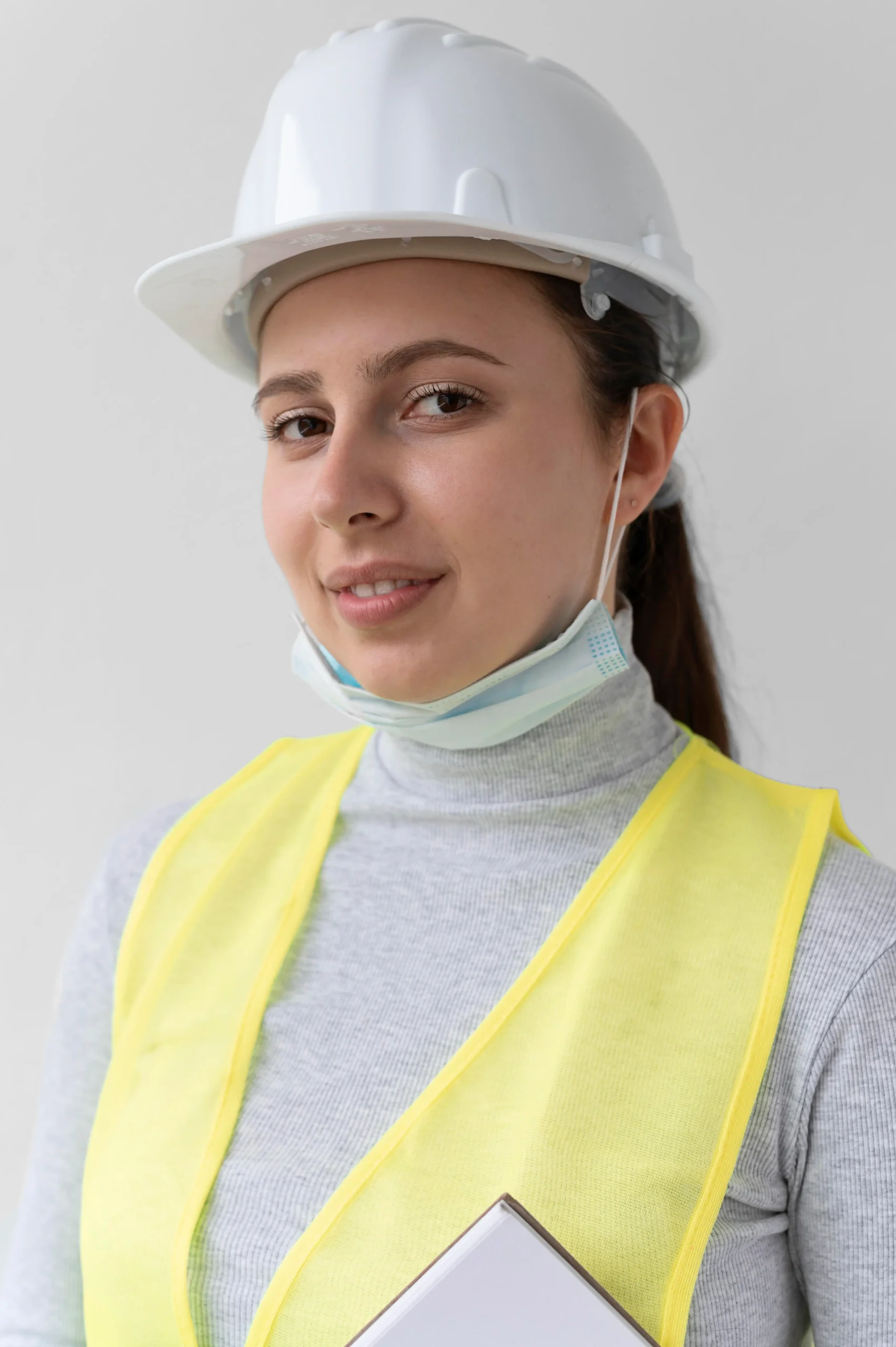 Mujer con casco blanco y chaleco de seguridad amarillo, cubrebocas bajado, sosteniendo una carpeta, sobre fondo neutro