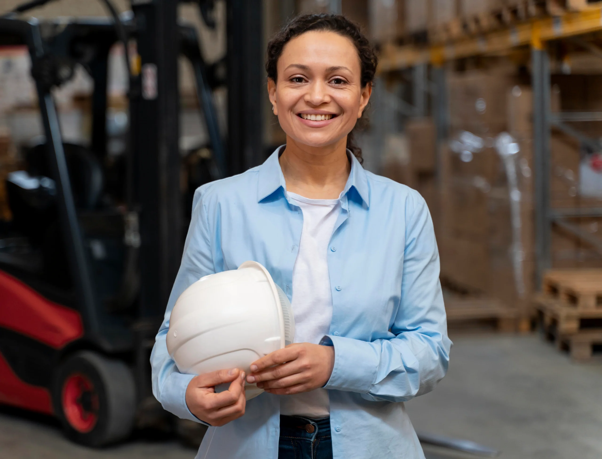 Mujer sonriente con camisa azul claro sosteniendo un casco blanco, de pie frente a montacargas y racks con mercancía envuelta en playo