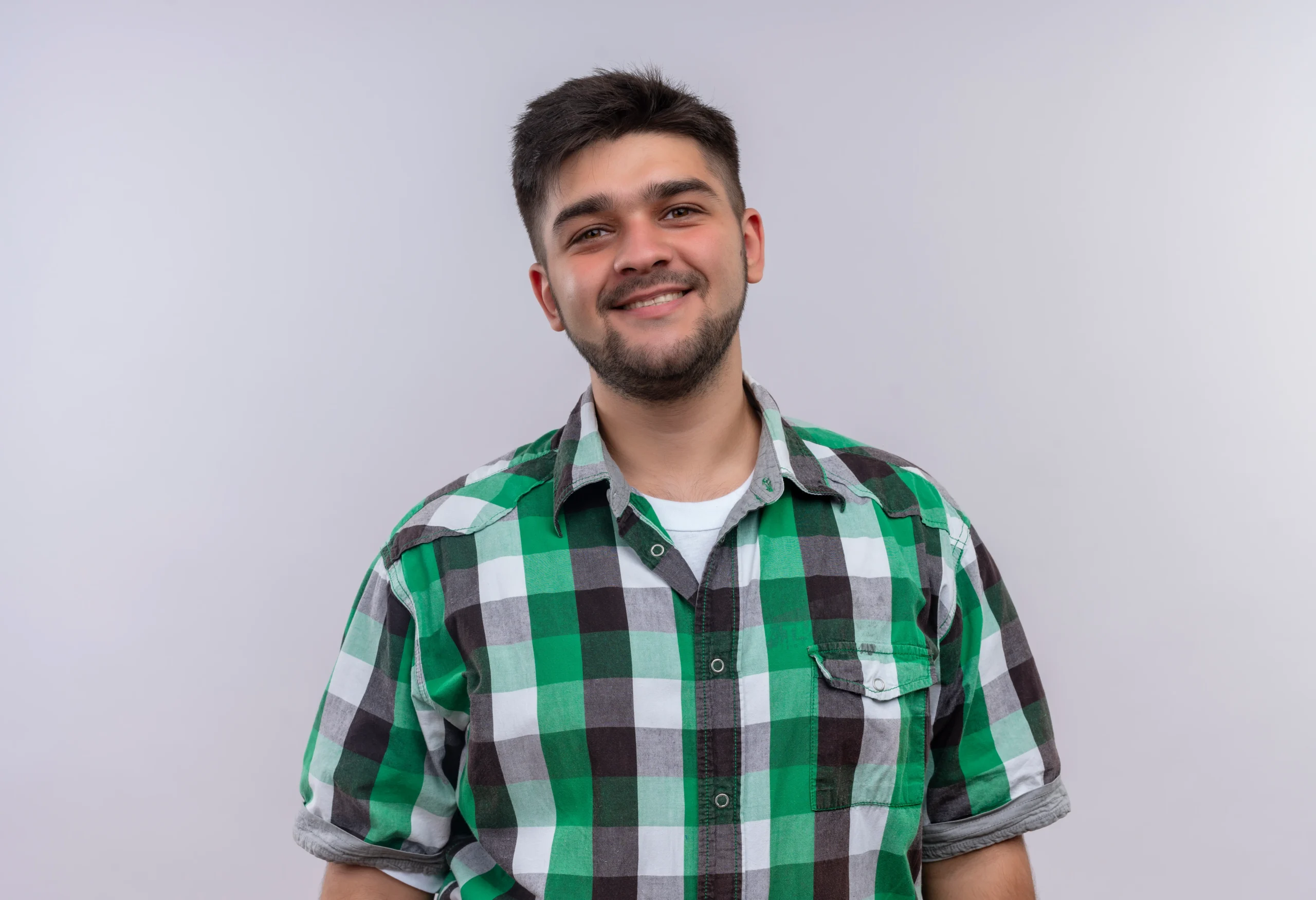 Hombre joven sonriente con barba, vistiendo camisa a cuadros verde y negro, sobre fondo blanco liso